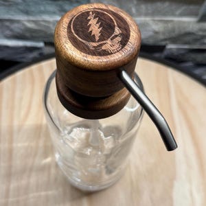 May include: A clear glass soap dispenser with a wooden top featuring a carved skull and lightning bolt design. The dispenser has a silver-colored pump and is designed for liquid soap or lotion. The dispenser is on a light-colored surface.