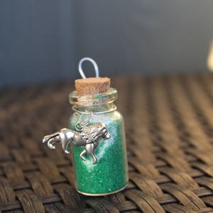 May include: A small glass bottle with a cork top filled with green glitter and a silver charm of a horse. The bottle is sitting on a woven wicker surface.