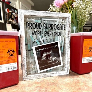 Op de afbeelding: Een wit houten frame met een glazen voorkant met een verzameling spuiten en een zwart-witte echografie afbeelding. Het frame heeft de tekst "PROUD SURROGATE WORTH EVERY SHOT" erop gedrukt. Twee rode biohazard containers zijn zichtbaar aan weerszijden van het frame.