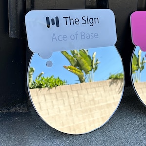 May include: A round, reflective mirror with the text "The Sign Ace of Base" on a white tag. The mirror reflects a blue sky, green plants, and a tan brick wall. The mirror is attached to a dark surface.