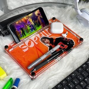 May include: A clear acrylic tray with an orange insert featuring a photo of a person and the word "SOPAIR". The tray holds a smartphone displaying a concert, a pair of white earbuds, a silver Sharpie, and a black pen. The tray is on a white fluffy surface.