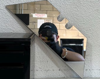 May include: A modern, geometric mirror with a unique shape and three curved cutouts at the top. The mirror is made of polished metal and reflects the surrounding environment. It is mounted on a white wall.