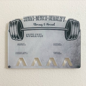 Peut inclure: Un tableau en acrylique gris et blanc avec un graphique de barre d'haltères noir et blanc. Le tableau comporte des espaces pour écrire les chiffres d'haltérophilie pour le squat, le soulevé de terre, le développé couché et le total. Le texte sur le tableau indique "SQUAT-BENCH-DEADLIFT Strong & Proud ATHLETE:"