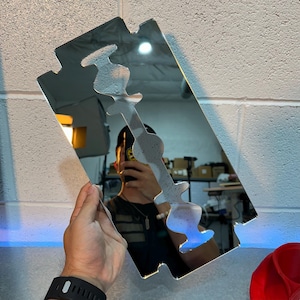 May include: A silver-colored mirror in the shape of a razor blade, held by a person. The mirror has a decorative cutout design. The background shows a white brick wall and a red object.