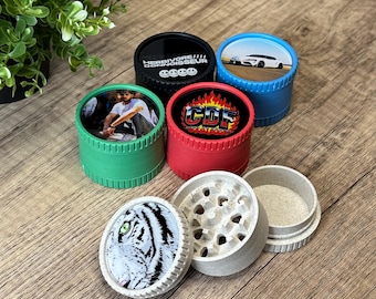 May include: A collection of colorful herb grinders with various designs. The grinders are in green, red, blue, and black. One grinder features a tiger's face, and others have images and text. The grinders are made of metal and plastic.