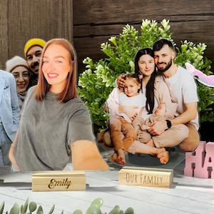 May include: Acrylic photo cutouts of people, including a family and an individual, with wooden nameplates that read "Emily" and "Our Family." The cutouts are placed in front of a green plant and a dark wood background.