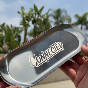 May include: A shiny, oval-shaped, silver metal tray with the word "Connected" in white script. The tray is held in front of a blurred background of palm trees and a blue sky.