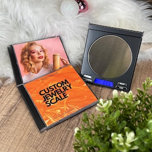 May include: A black digital jewelry scale with a silver weighing surface and a blue digital display showing 0.06. Beside it is a CD case with the text "CUSTOM JEWELRY SCALE" on an orange background and a photo of a woman.