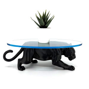 May include: A black panther sculpture serving as a base for an oval glass tabletop with a blue edge. A small green succulent plant sits in a white pot on the center of the glass surface. The panther is in a prowling stance with its tail curved.
