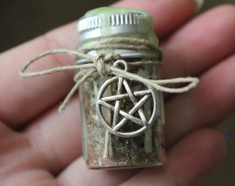 May include: A small glass jar filled with dried herbs and spices, sealed with a silver lid and green wax. A twine bow and a silver pentacle charm adorn the jar. The jar is held in a person's hand.