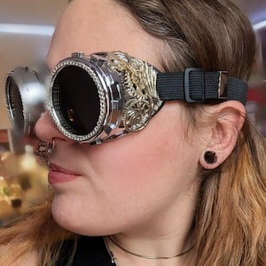 May include: Steampunk goggles featuring silver and gold metal detailing. The goggles have dark lenses and an adjustable black elastic strap. The design incorporates intricate gear and filigree patterns, creating a vintage, industrial aesthetic.