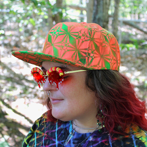 Peut inclure: Une personne portant une casquette de baseball orange vif et verte à motifs et des lunettes de soleil rouges et dorées uniques avec des accents rouges dégoulinants. La personne porte une chemise tie-dye colorée, avec des cheveux roux.