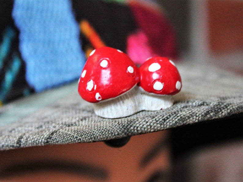 May include: Two red and white ceramic mushrooms with white polka dots. The mushrooms are sitting on a gray textured surface.