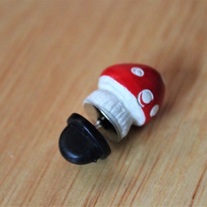 May include: A red and white mushroom shaped pin with a black pin back.