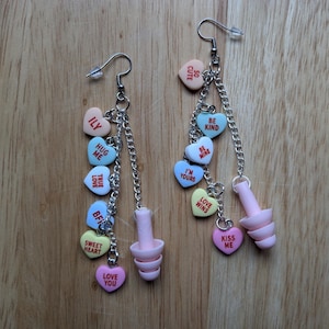 May include: A pair of earrings with pink, blue, and yellow heart-shaped charms. Each charm has a different message, such as "ILY", "Hug Me", "True Love", "Be Mine", "I'm Yours", "Love Wins", "Kiss Me", "So Cute", and "Be Kind".