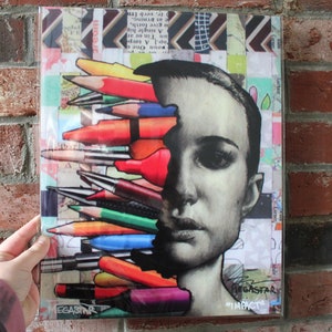 Puede incluir: Una obra de arte de collage colorida que presenta el rostro de una mujer parcialmente oscurecido por una colección de lápices y bolígrafos. La obra de arte se encuentra sobre un fondo de papel estampado e incluye el texto "MEGASTAR" y "#IMPACT".