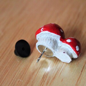 May include: A red and white mushroom shaped pin with a black plastic backing. The pin is attached to a silver metal pin.