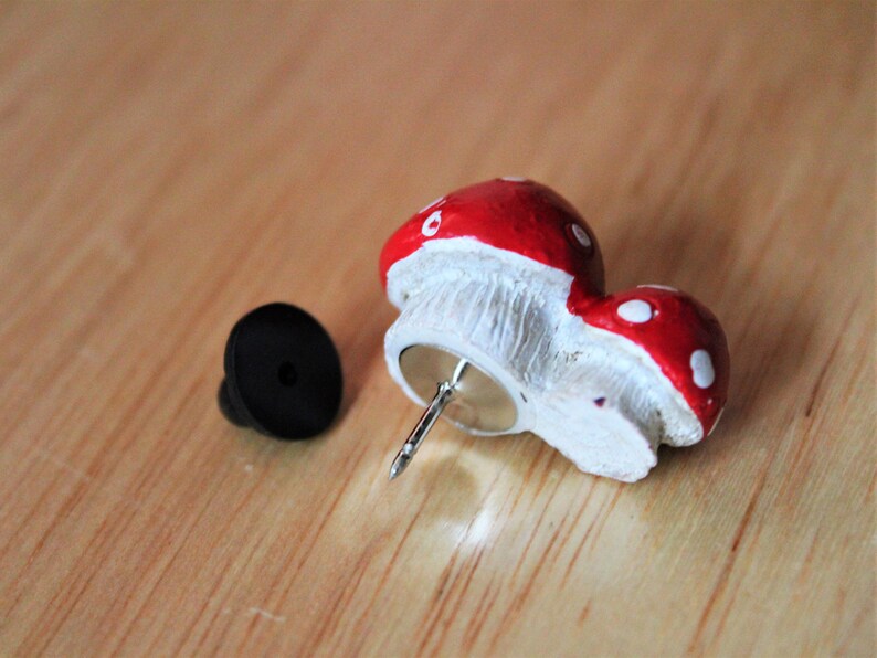 May include: A red and white mushroom shaped pin with a black plastic backing. The pin is attached to a silver metal pin.
