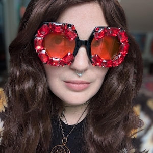 May include: Black-framed sunglasses with large, red, jewel-like embellishments around the lenses. The lenses are a warm orange color. The sunglasses have a bold, geometric shape, creating a statement accessory.