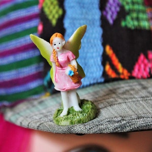 May include: A small fairy figurine in a pink dress, with orange braided hair and green wings, carrying a basket of blue flowers. The fairy stands on a patch of green grass, on a patterned hat with a colourful textile design.