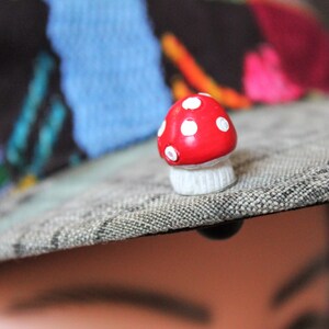 May include: A red and white mushroom-shaped pin with white polka dots. The pin is attached to a gray fabric surface.