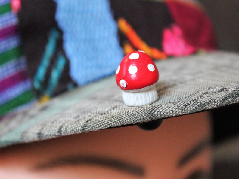 May include: A red and white mushroom-shaped pin with white polka dots. The pin is attached to a gray fabric surface.