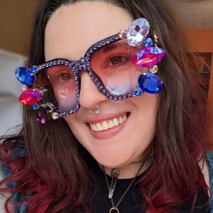 Peut inclure: Lunettes de soleil carrées surdimensionnées avec une monture violette et des verres roses. La monture est ornée de nombreux gros strass colorés dans les tons bleu, rose et transparent. Les lunettes ont une esthétique audacieuse et glamour.