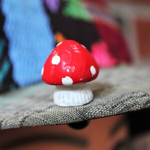 May include: A small red and white mushroom figurine with white polka dots. The mushroom is sitting on a textured gray surface.