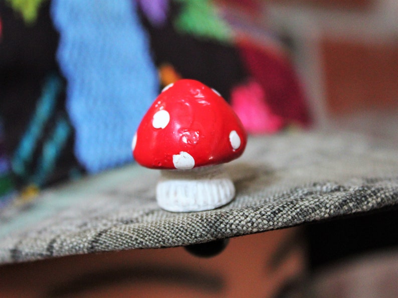 May include: A small red and white mushroom figurine with white polka dots. The mushroom is sitting on a textured gray surface.