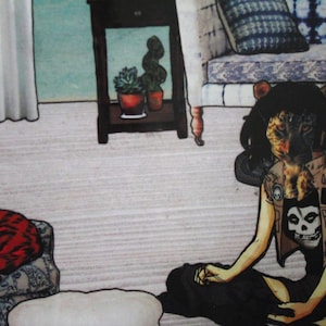 May include: A woman with a leopard head sits on the floor with her legs crossed. She is wearing a black and brown vest with a skull design and a black shirt. She is sitting in front of a couch with a blue and white patterned cushion.