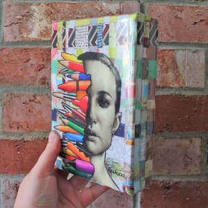 May include: A colorful collage of colored pencils and a woman's face on a small canvas. The canvas is covered in colorful paper scraps and has a glossy finish.