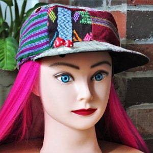 May include: A colorful patchwork baseball cap with a black brim and red mushroom embellishments. The cap is worn on a mannequin with bright pink hair.