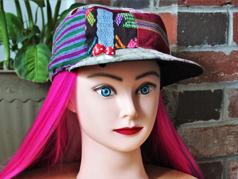 May include: A colorful patchwork baseball cap with a black brim and red mushroom embellishments. The cap is worn on a mannequin with bright pink hair.