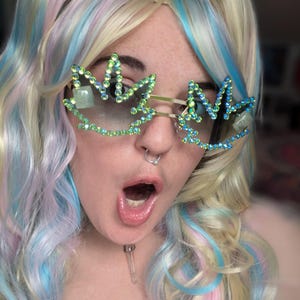 May include: A close-up of a person wearing novelty sunglasses. The sunglasses have a gold frame and are shaped like cannabis leaves, embellished with green and blue rhinestones. The person has blonde and pastel-colored hair.