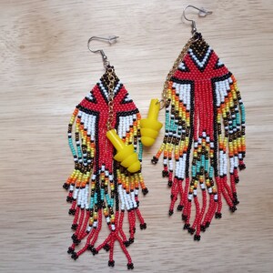 May include: A pair of long, beaded earrings with a geometric pattern in red, white, black, yellow, and turquoise. Each earring has a yellow earplug charm hanging from it.