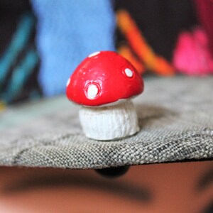 May include: A small red and white mushroom figurine with white polka dots on the top. The mushroom is sitting on a gray textured surface.