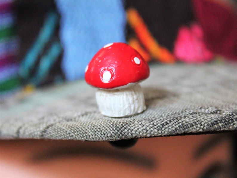 May include: A small red and white mushroom figurine with white polka dots on the top. The mushroom is sitting on a gray textured surface.