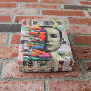 May include: A colorful collage artwork featuring a woman's face with a variety of colored pencils and markers layered over her face. The artwork is mounted on a canvas and has a textured background. The text "MEGAFAR" is visible in the lower right corner of the artwork.