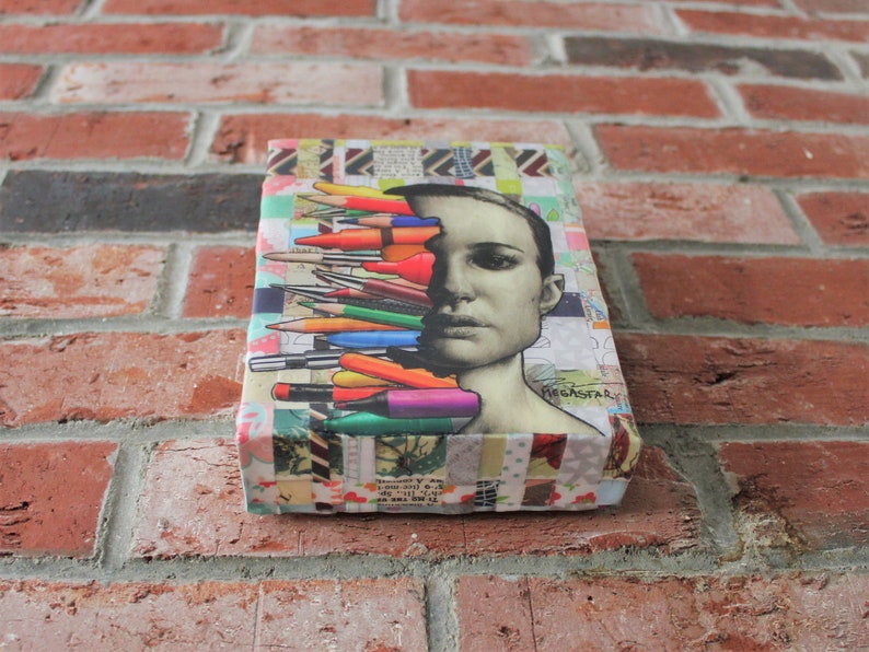 May include: A colorful collage artwork featuring a woman's face with a variety of colored pencils and markers layered over her face. The artwork is mounted on a canvas and has a textured background. The text "MEGAFAR" is visible in the lower right corner of the artwork.