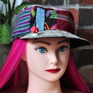 May include: A colorful patchwork fabric cap with a mushroom pin. The cap has a flat brim and is made of a variety of fabrics in different colors and patterns.