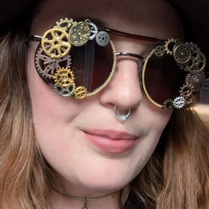 May include: Brown steampunk sunglasses with decorative metal gears and cogs around the lenses. The glasses have a dark tint and a gold-colored frame. The gears are in various sizes and metallic colors.