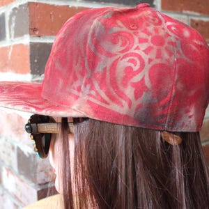 May include: A red and white baseball cap with a faded floral pattern. The cap is being worn by a person with long brown hair.