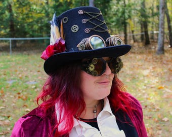 May include: Black steampunk top hat with brass gears, chains, and goggles. The hat is decorated with a feather and a red flower. The goggles have round lenses and intricate gear detailing. The person is wearing a burgundy velvet jacket.