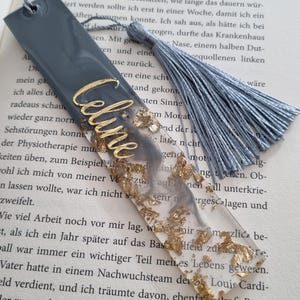 May include: A rectangular resin bookmark featuring a gray and clear marbled design with embedded gold leaf flakes. The name Celine is written in gold script lettering on the surface. A gray silk tassel is attached to the top hole of the bookmark.