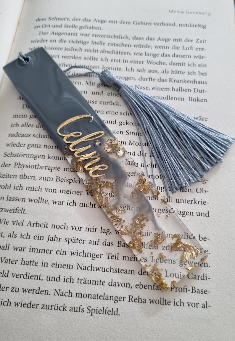 May include: A rectangular resin bookmark featuring a gray and clear marbled design with embedded gold leaf flakes. The name Celine is written in gold script lettering on the surface. A gray silk tassel is attached to the top hole of the bookmark.