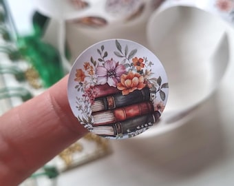 May include: A circular sticker featuring a stack of three books in shades of red, black, and brown, topped with a colorful arrangement of orange, pink, and white flowers with green leaves and stems, set against a plain white background.