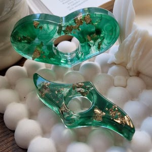 May include: Two translucent green resin objects with embedded gold leaf flakes. The top object is heart-shaped with a central circular opening, and the bottom object is a curved, elongated shape with a central circular opening.
