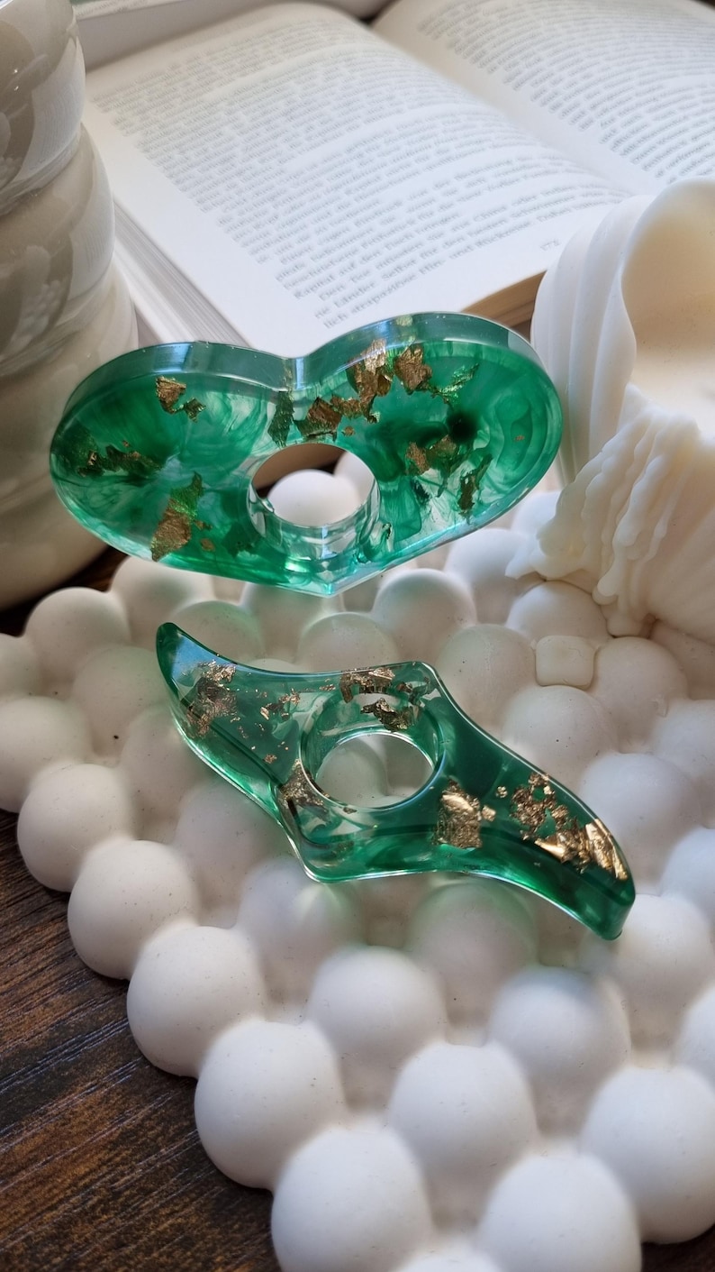 May include: Two translucent green resin objects with embedded gold leaf flakes. The top object is heart-shaped with a central circular opening, and the bottom object is a curved, elongated shape with a central circular opening.