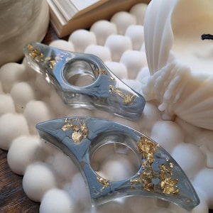 May include: Two crescent-shaped resin finger guards in a translucent gray finish with embedded gold leaf flakes. Each piece features a central circular opening designed to fit a finger, showcasing a smooth, polished surface texture.