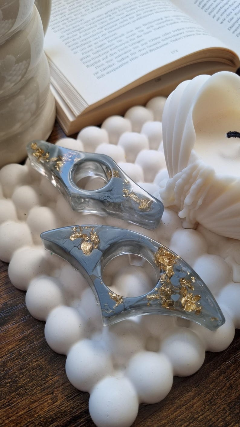 May include: Two crescent-shaped resin finger guards in a translucent gray finish with embedded gold leaf flakes. Each piece features a central circular opening designed to fit a finger, showcasing a smooth, polished surface texture.